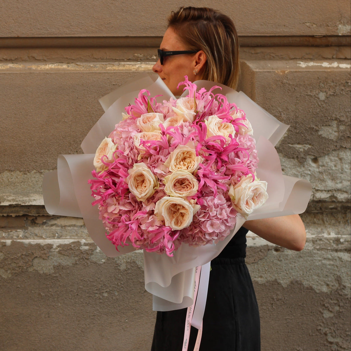A woman in a black dress and sunglasses holds a large bouquet of pink and cream roses wrapped in white tissue paper, standing against a textured, peeling-stone wall.