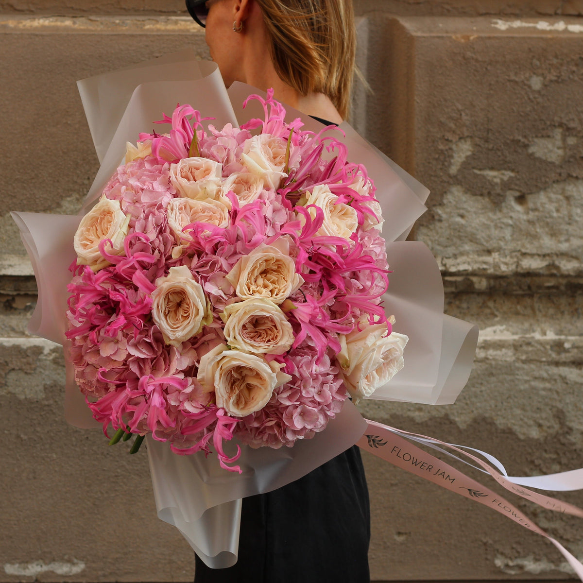 A person from behind holding a large bouquet of pink and cream roses wrapped in translucent white paper, standing against a textured stone wall.