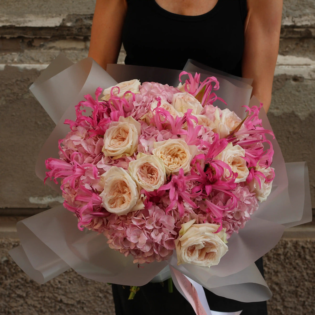 A large, vibrant flower bouquet featuring pink hydrangeas, blush pink roses, and bright pink lilies wrapped in translucent white paper, held by a person wearing a black top against a rustic stone wall.