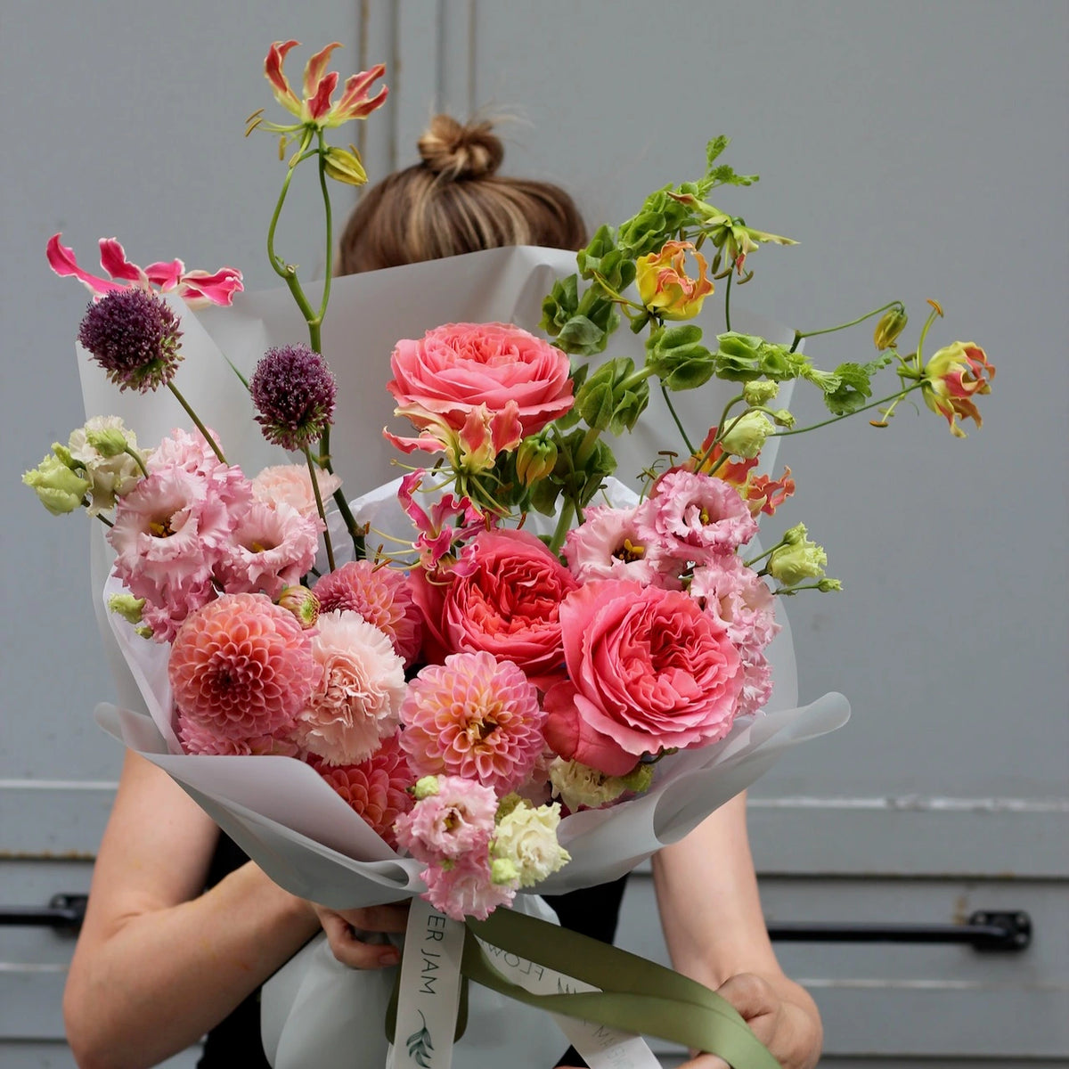 A woman holiding a bouquet of pink colored flowers on a grey background wrapped in a neutral paper. 
