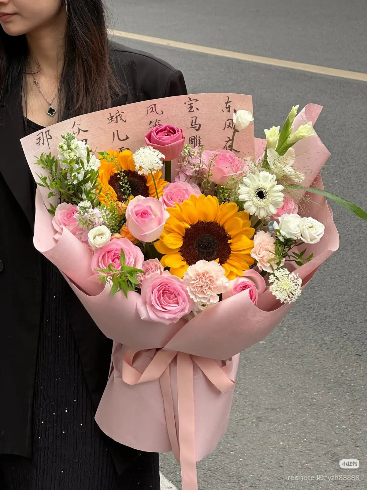 A woman holding a bouquet with sunflower and pink roses and white flowers, inside a pink envelope.