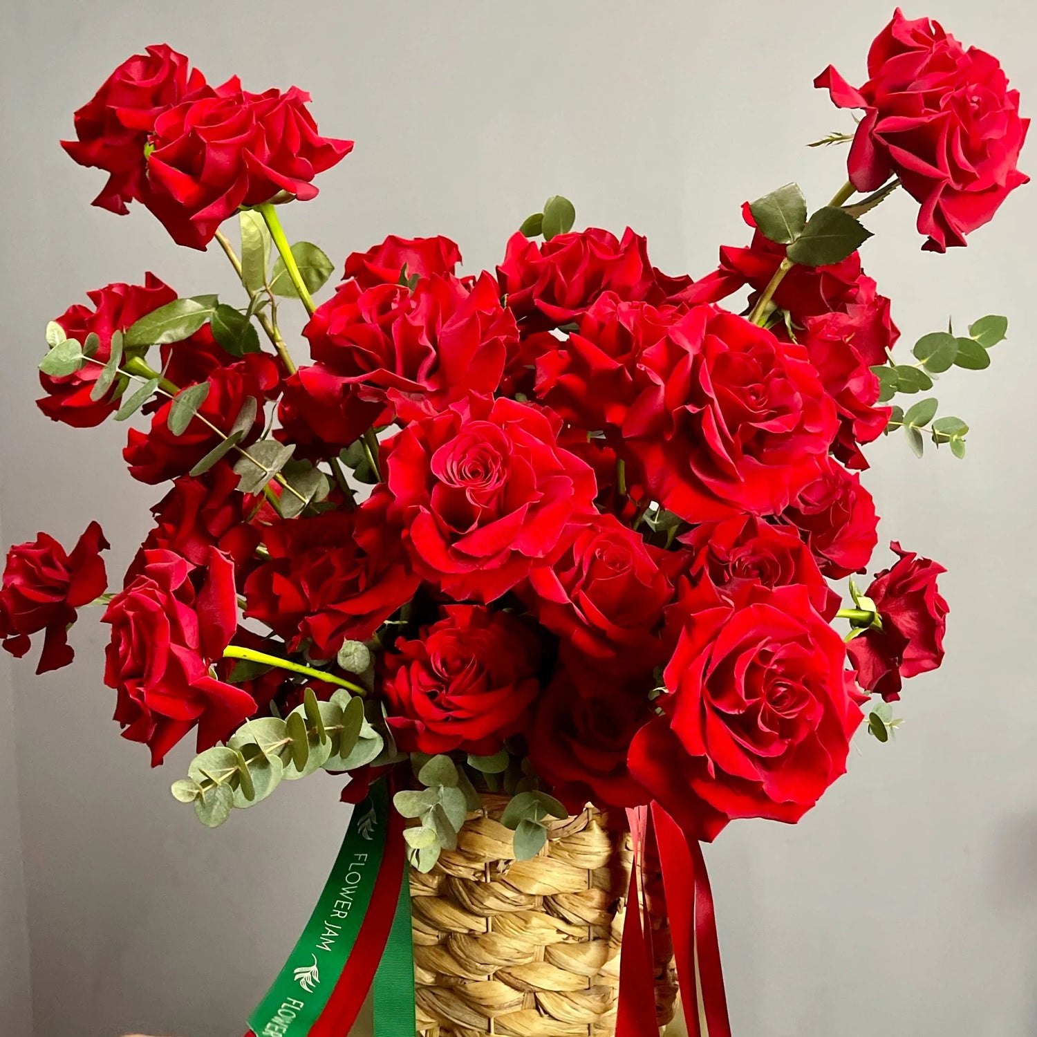Bouquet of red roses in a woven basket with a visible brand tag against a plain background