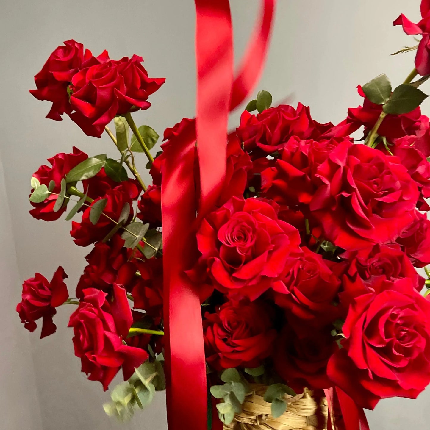 Bouquet of red roses with a red ribbon in a woven basket against a neutral background